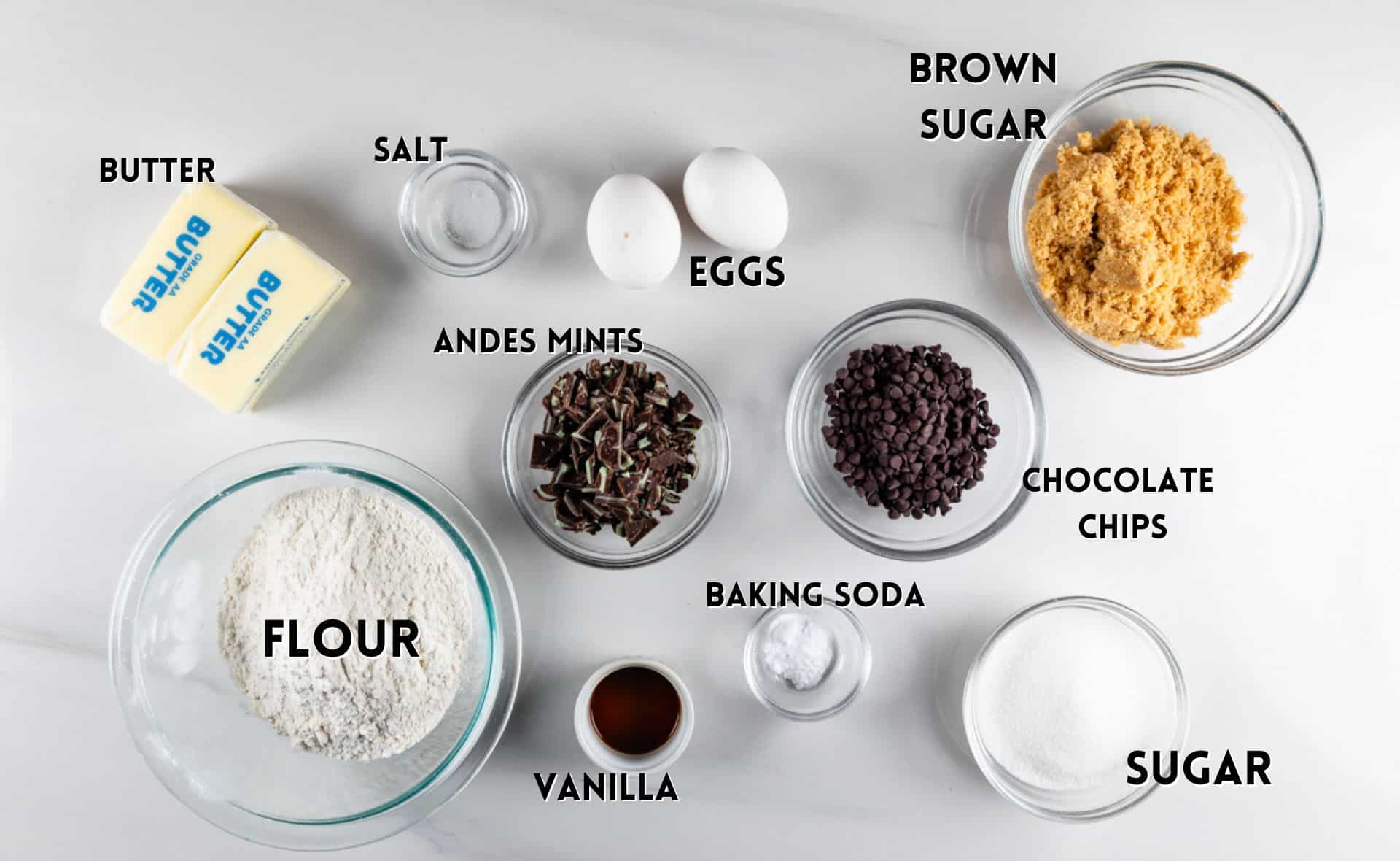 A top-down view of baking ingredients labeled on a white surface: butter, eggs, brown sugar, chocolate chips, sugar, baking soda, vanilla, Andes mints, flour, and salt. Each ingredient is in a separate container or package.