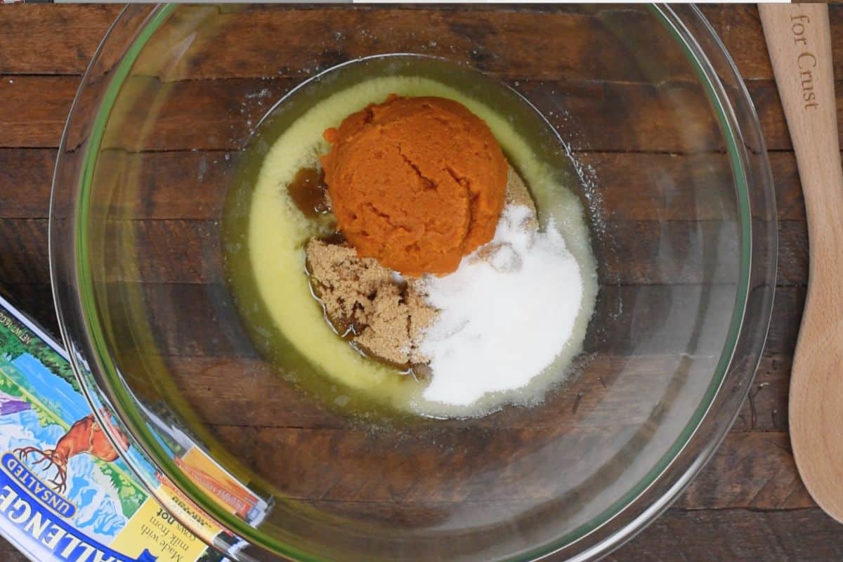 A glass bowl on a wooden table contains pumpkin puree, brown sugar, white sugar, and melted butter. A wooden spoon with engraved text and a colorful magazine are partially visible on the table.