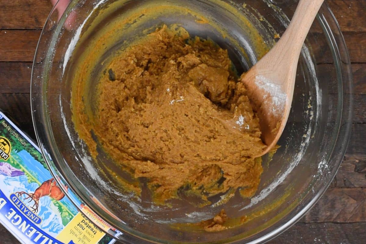 A glass bowl filled with a brown cookie dough mixture. A wooden spoon rests in the bowl. Next to the bowl, part of a package featuring colorful graphics is visible on a wooden surface.