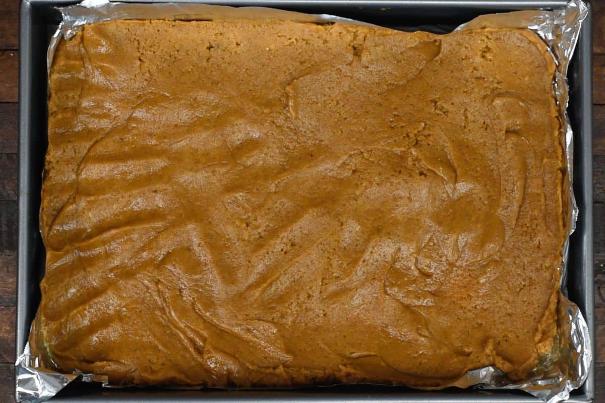 A rectangular baking pan lined with foil is filled with a smooth, golden-brown batter, ready to be baked. The batters surface shows swirled patterns and has a creamy texture.