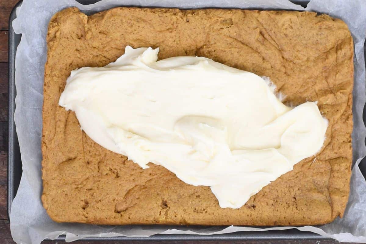 A large rectangular pumpkin cake on a baking tray with parchment paper. A generous dollop of cream cheese frosting is spread across the center, ready to be evenly distributed.