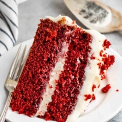 A slice of red velvet cake with creamy white frosting sits on a white plate. A fork is placed next to the cake, and a striped napkin is partially visible in the background.