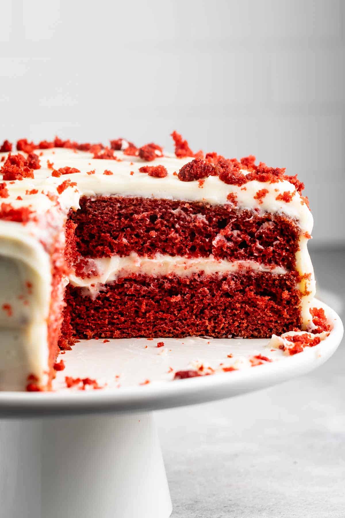 A red velvet cake with cream cheese frosting is displayed on a white cake stand. A slice is cut out, revealing the cakes two layers and smooth frosting. Crumbs are scattered on top and around the cake.