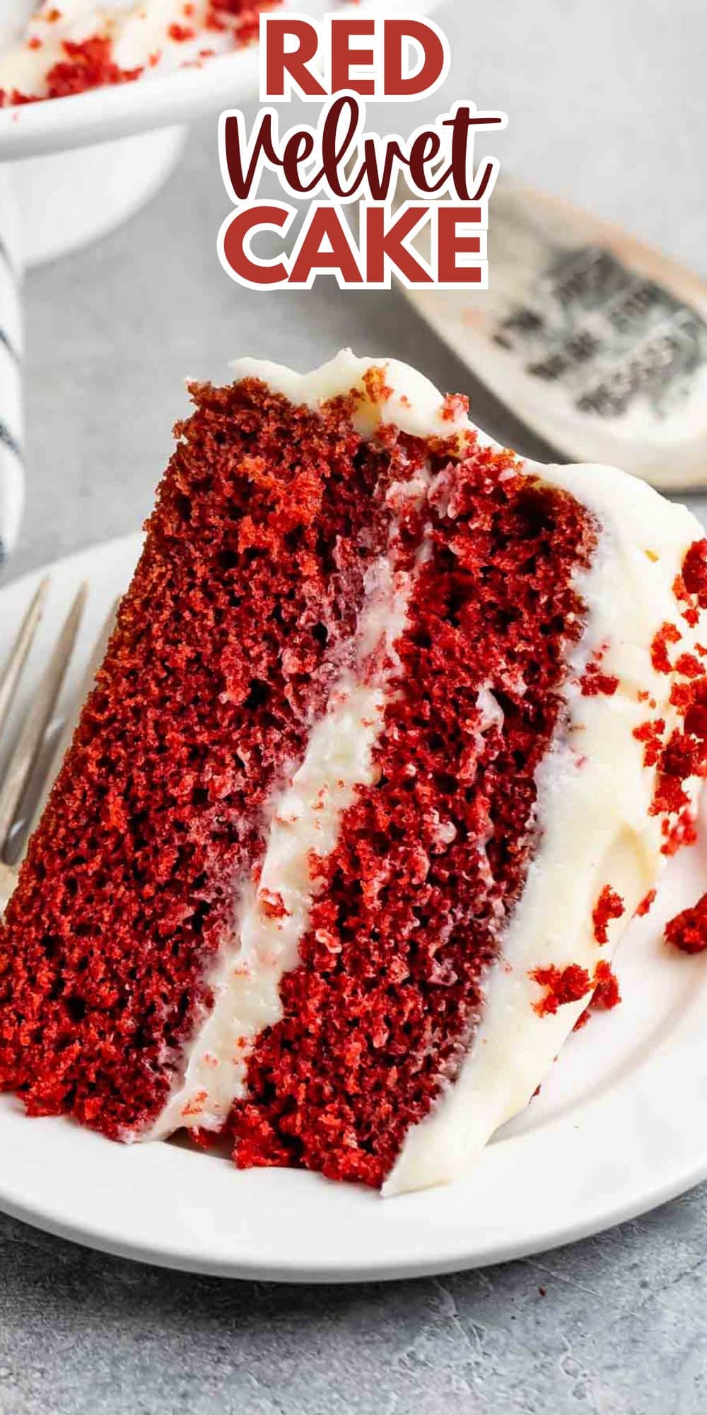 A slice of red velvet cake with cream cheese frosting on a white plate. The cake has two layers, and there are red crumbs scattered on the frosting. A fork is placed beside the slice. Text above reads Red Velvet Cake.