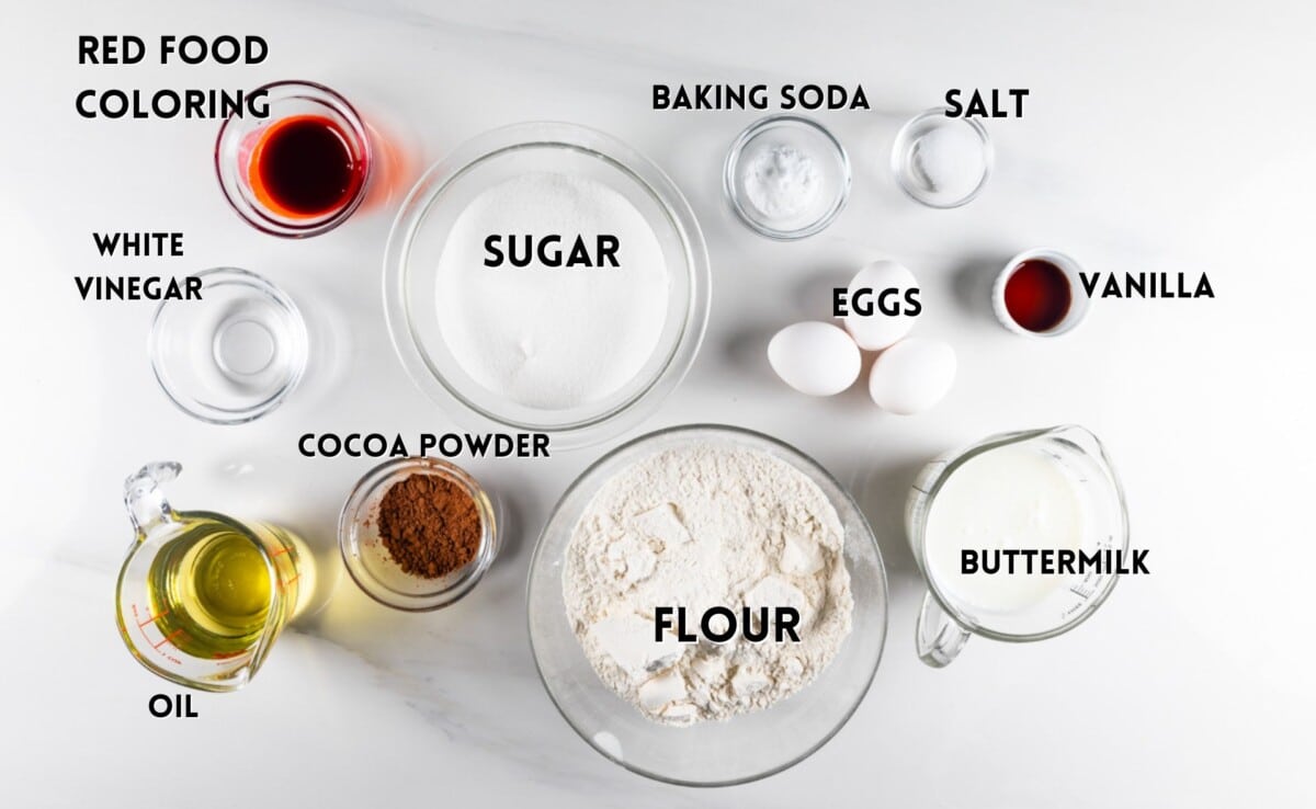 Top view of baking ingredients labeled on a white surface: red food coloring, white vinegar, cocoa powder, oil, sugar, flour, baking soda, salt, eggs, vanilla, and buttermilk in separate containers.