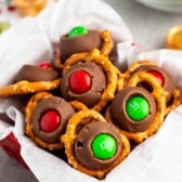 A festive bowl of sweet treats featuring chocolate rosettes topped with red and green candies, nestled on circular pretzels. The background shows a bowl with more red and green candies and foil-wrapped chocolates, creating a holiday atmosphere.