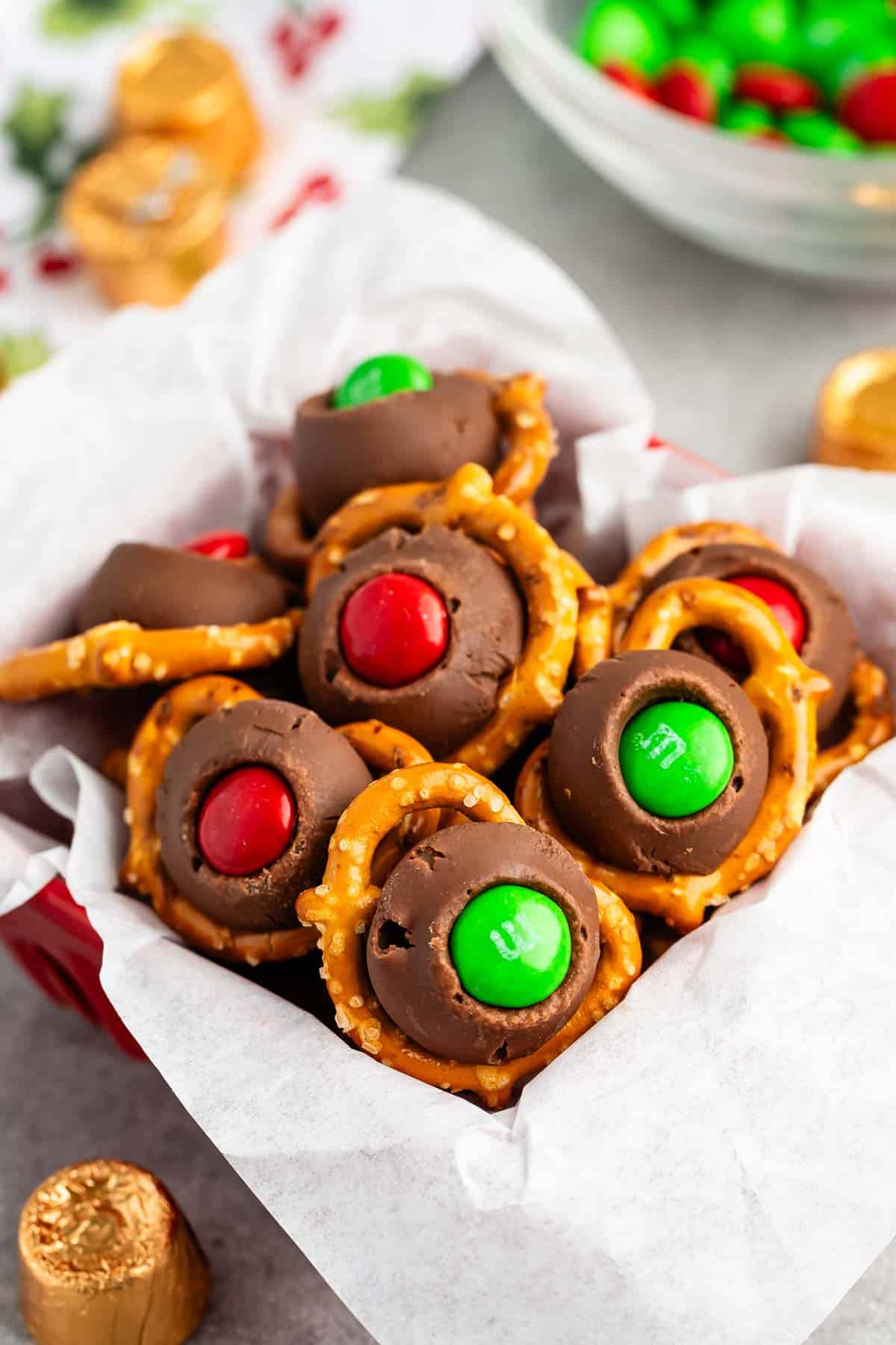 A festive bowl of sweet treats featuring chocolate rosettes topped with red and green candies, nestled on circular pretzels. The background shows a bowl with more red and green candies and foil-wrapped chocolates, creating a holiday atmosphere.