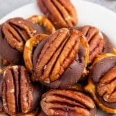 A plate of chocolate-covered pretzels topped with salted pecans. The treats are stacked on a white plate, showcasing a mix of smooth chocolate, crunchy pretzels, and whole pecans.