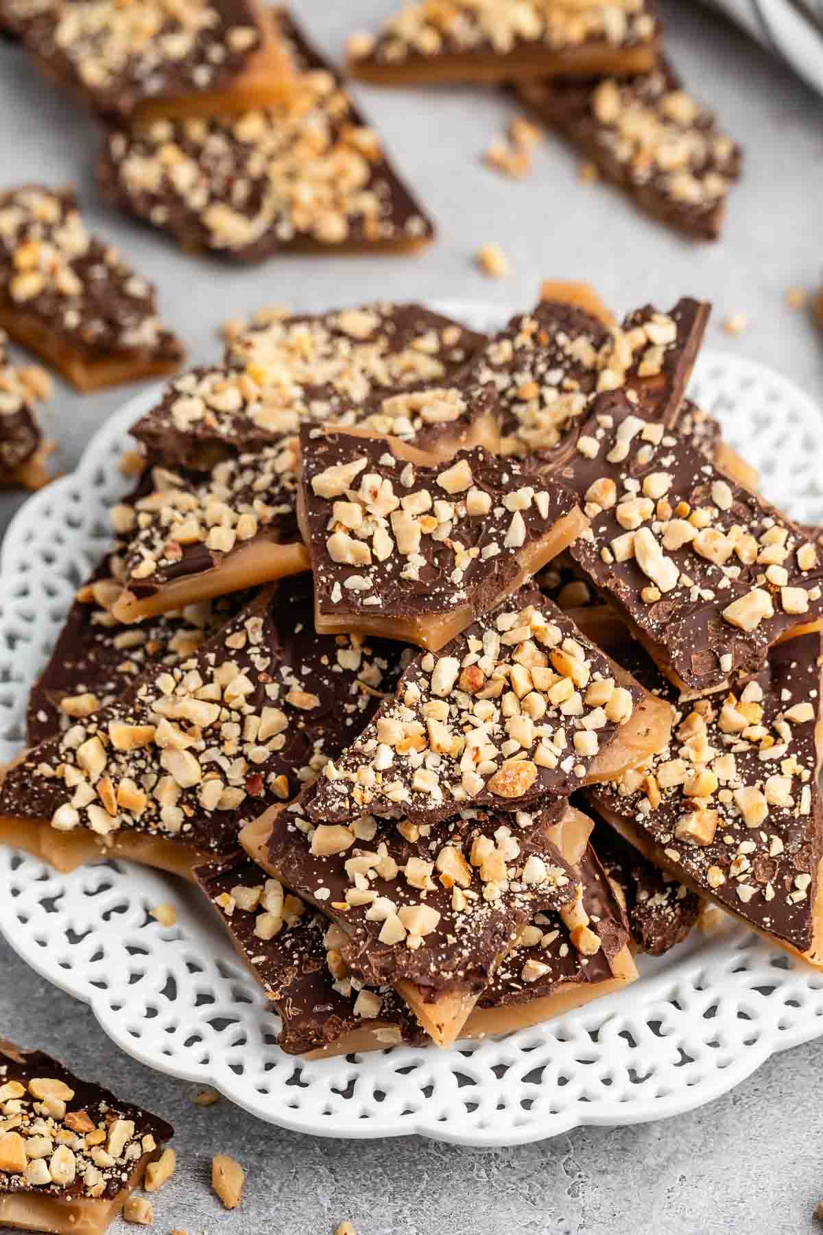 A white lace-edged plate piled with pieces of chocolate toffee, topped with chopped nuts. Additional pieces of toffee are scattered around the plate on a gray surface.