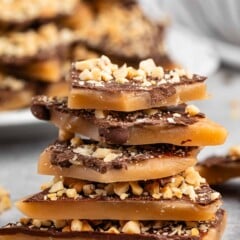 A stack of toffee pieces topped with chocolate and crushed nuts is placed on a gray surface. More toffee pieces are blurred in the background, giving a delicious and homemade dessert vibe.