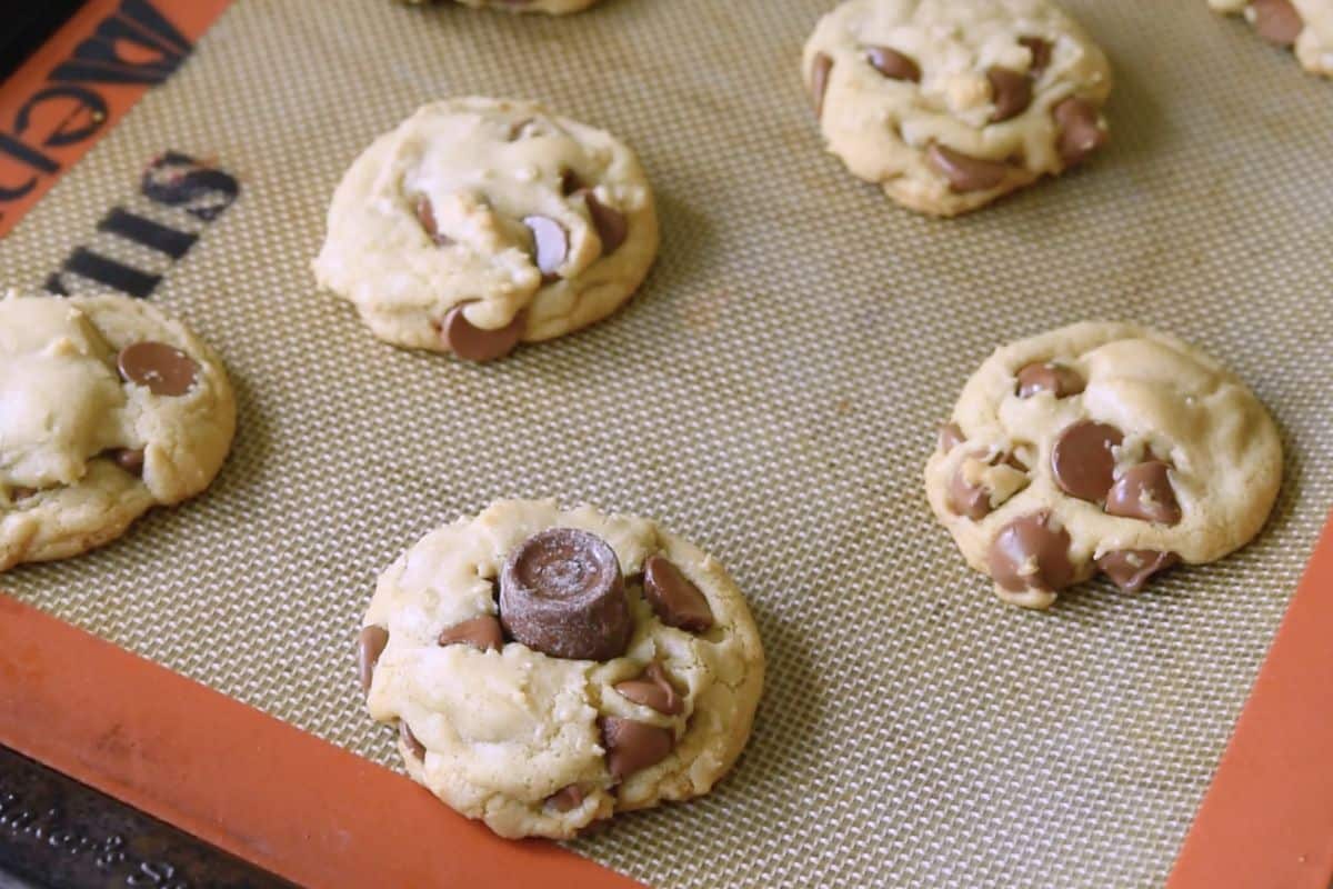 A baking tray with six freshly baked chocolate chip cookies on a silicone mat. One cookie has a chocolate candy piece in the center. The cookies are golden brown and evenly spaced on the mat.