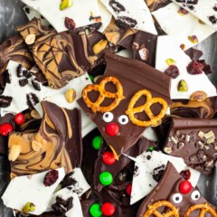 A festive assortment of chocolate bark featuring white and dark chocolate pieces. Toppings include pretzels, candy eyes, M&Ms, nuts, dried cranberries, and cookie crumbs arranged creatively on a tray.