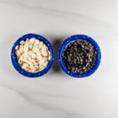 Two blue bowls on a light countertop, one filled with white chocolate chips and the other with dark chocolate chips.