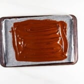 A baking tray lined with parchment paper holds a layer of smooth, melted chocolate spread evenly across its surface.