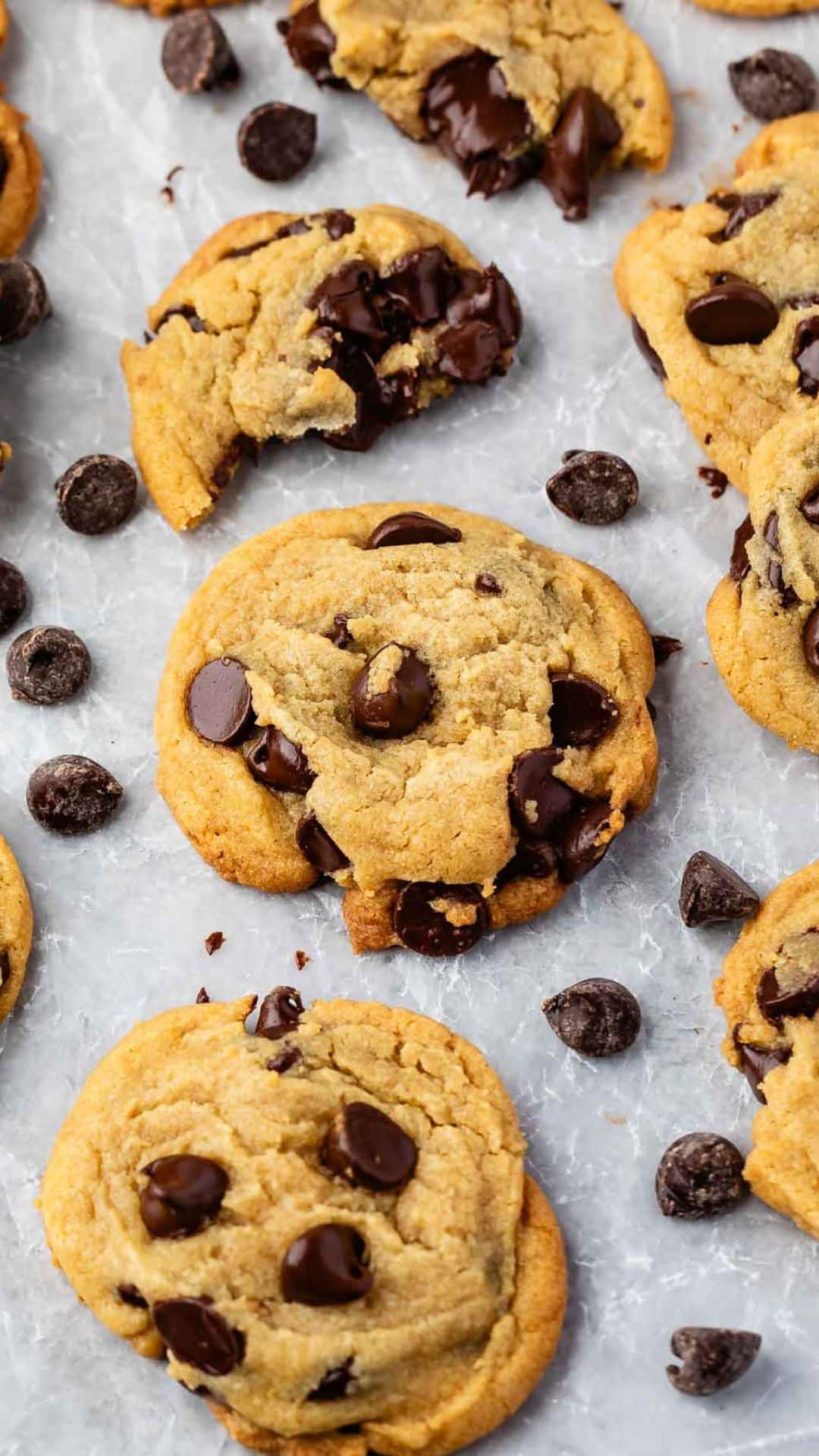 A close-up of chocolate chip cookies and scattered chocolate chips on a textured surface. The cookies are golden brown with visible chunks of chocolate, creating an appetizing display.