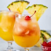 Two glasses of tropical cocktails garnished with pineapple slices and cherries. The drinks are orange with ice cubes, set on a light gray surface. A small bowl of cherries is visible in the background.