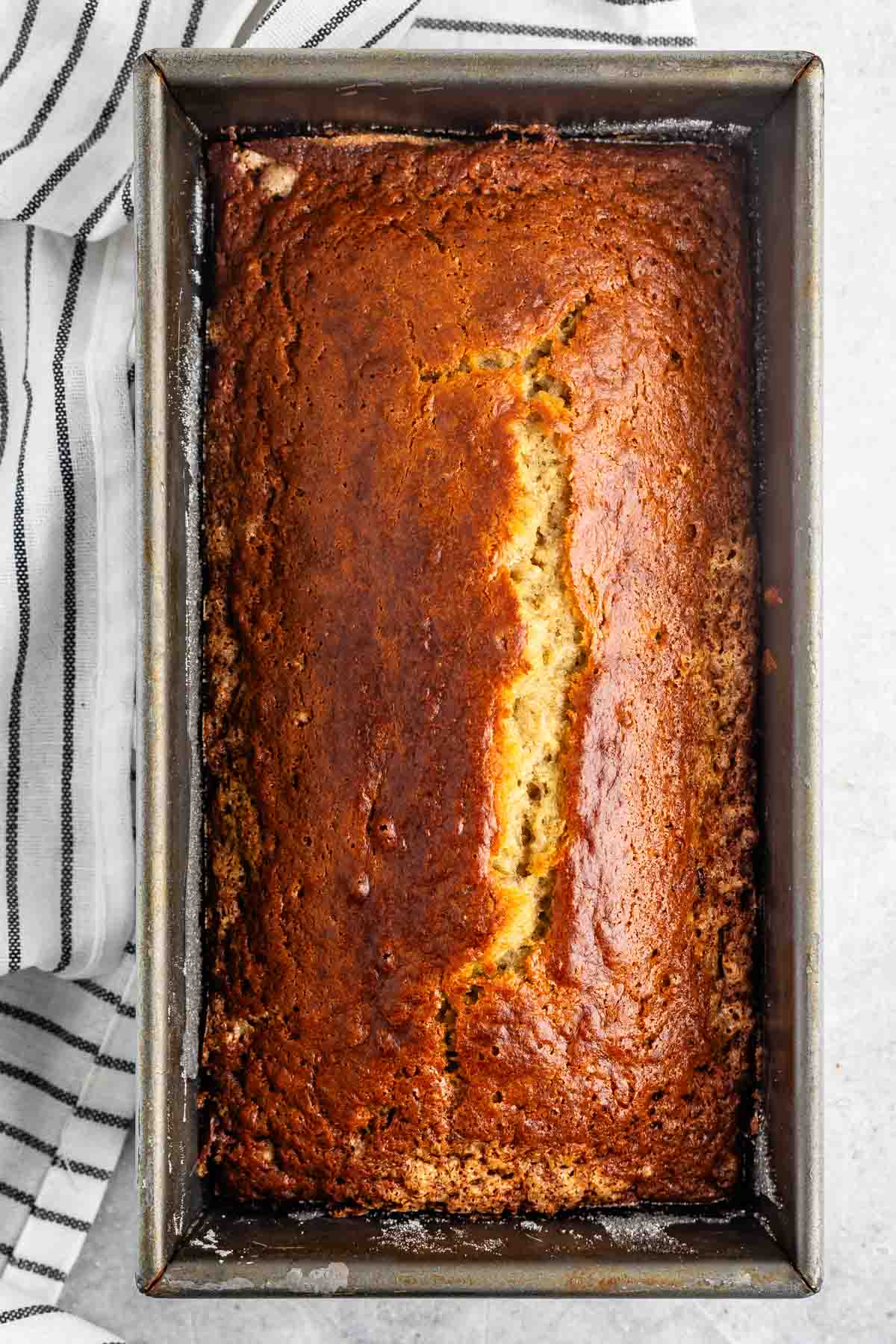 A freshly baked loaf of banana bread in a rectangular metal pan. The bread has a golden-brown crust with a slight crack down the middle, resting on a light surface with a striped cloth next to it.