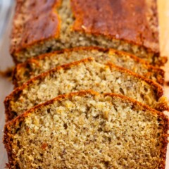 A close-up of a freshly baked banana bread loaf, partially sliced, with a golden-brown crust. The breads moist texture and small air pockets are visible, highlighting its homemade quality. It rests on a wooden cutting board.