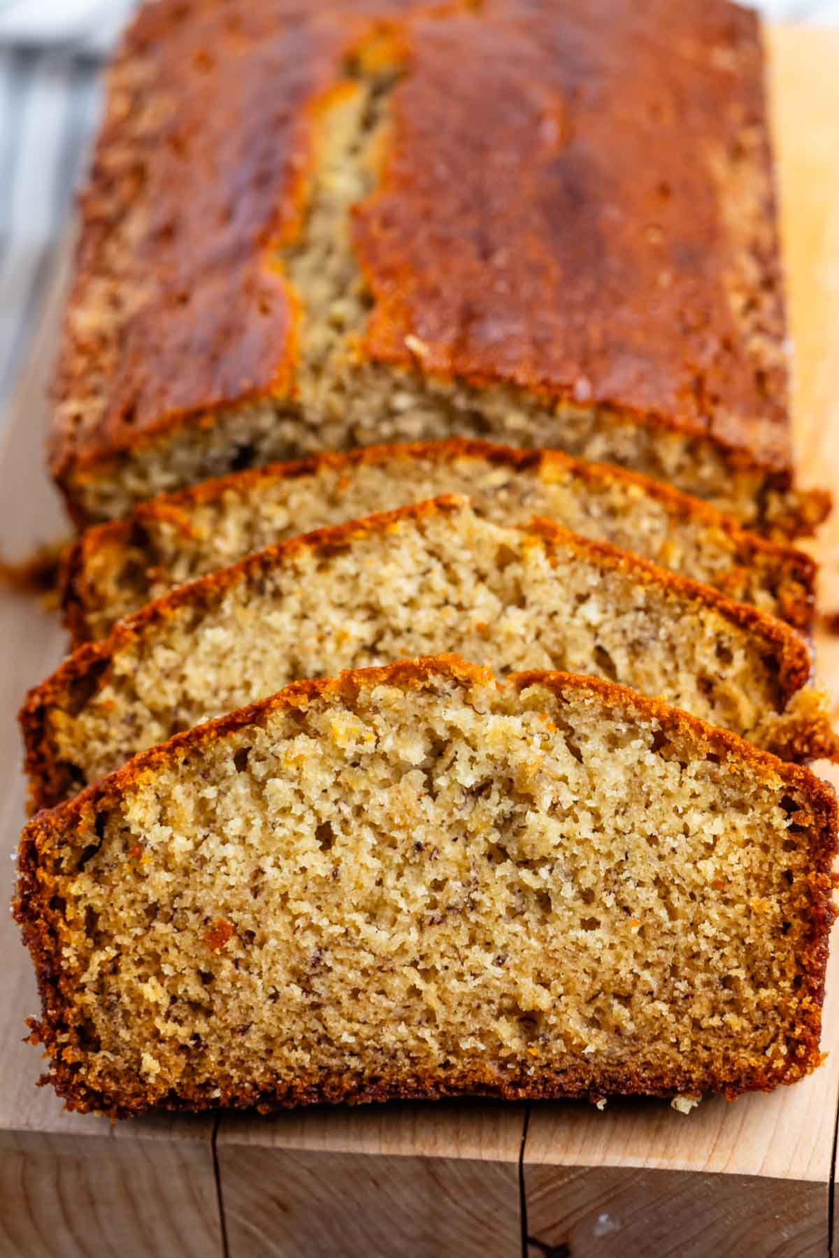 A close-up of a freshly baked banana bread loaf, partially sliced, with a golden-brown crust. The breads moist texture and small air pockets are visible, highlighting its homemade quality. It rests on a wooden cutting board.