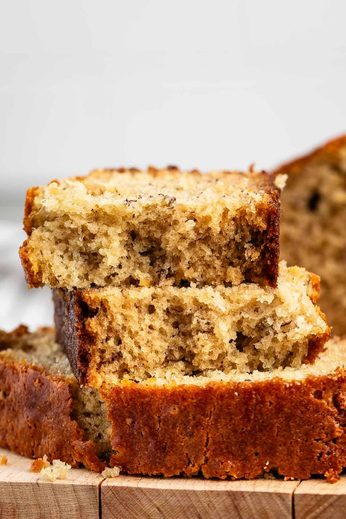 Close-up of slices of banana bread stacked on a wooden cutting board. The bread has a golden-brown crust and a moist, dense interior with visible banana flecks. More slices and the loaf are partially visible in the background.