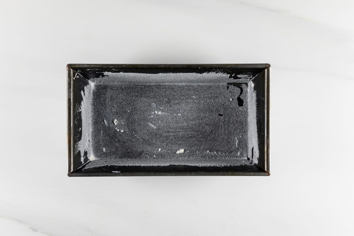 A rectangular baking pan with a light coating of flour or sugar. The surface appears slightly uneven, indicating previous use or preparation. The pan rests on a white marble background, viewed from above.