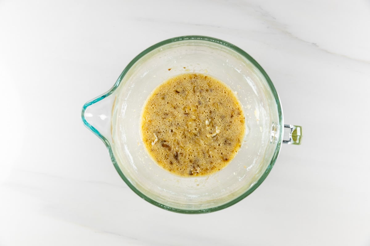 A glass mixing bowl with a frothy, golden-brown batter sits on a white marble surface, viewed from above. The batter contains visible bubbles and small bits, indicating preparation for baking or cooking.