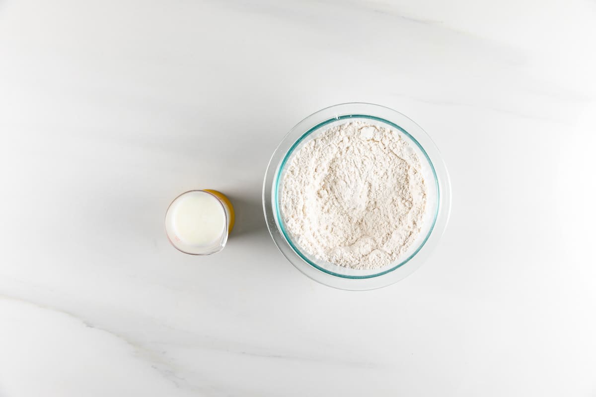 A glass bowl filled with flour and a small glass of milk are placed on a white marble surface. The bowl is on the right and the glass of milk is on the left.