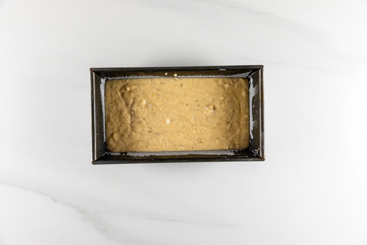 A loaf pan filled with banana bread batter placed on a white marble countertop, ready to be baked.