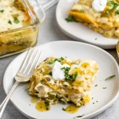 A close-up of two plates each with a serving of creamy chicken enchilada casserole topped with fresh cilantro and dollops of sour cream. A glass baking dish with more casserole is visible in the background. Forks are placed on the plates.