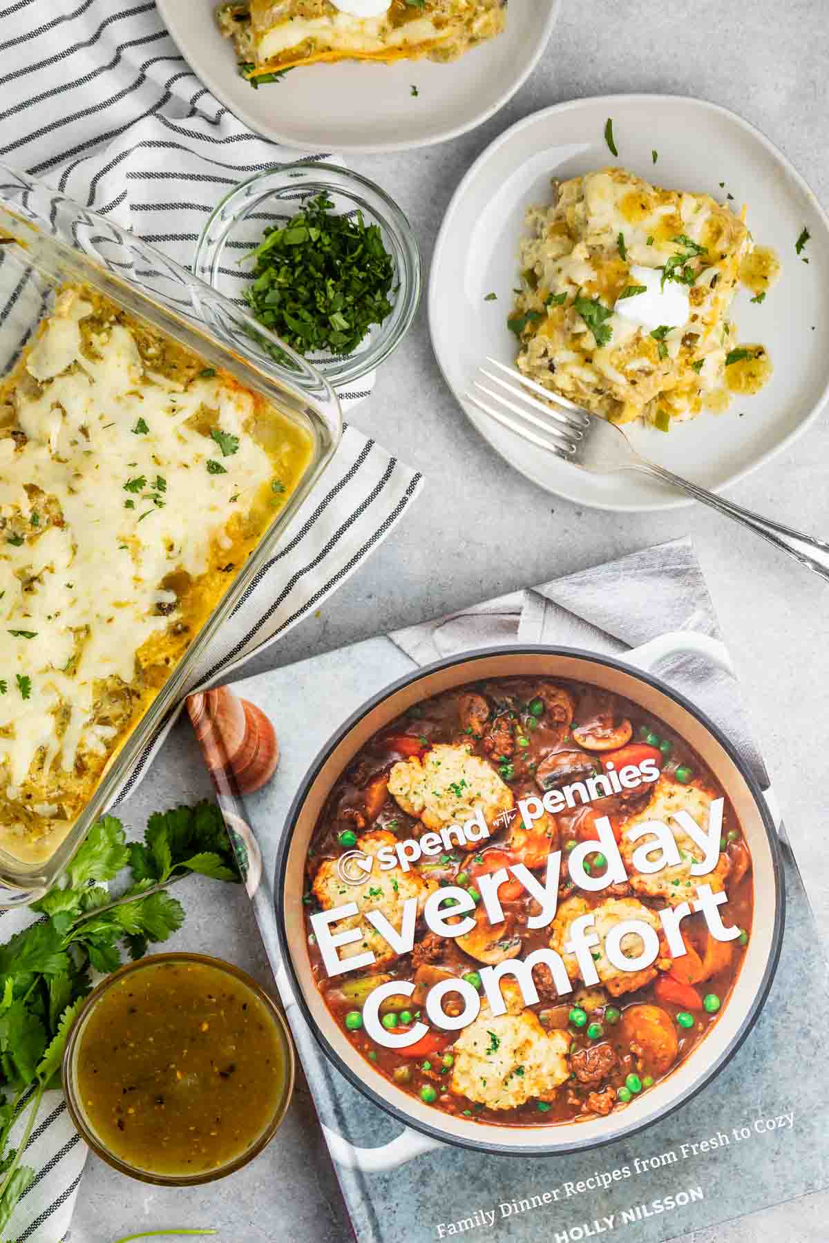 A cookbook titled Everyday Comfort lies on a table next to a serving of casserole on a plate. The casserole dish is partly full, with fresh herbs sprinkled on top. A small bowl of green herbs and a napkin are also visible.