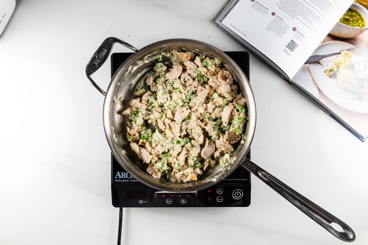 A pot of creamy pasta with chunks of chicken and broccoli is cooking on an induction stove. An open cookbook lies nearby, partially visible, with a leafy green dish pictured on one page.