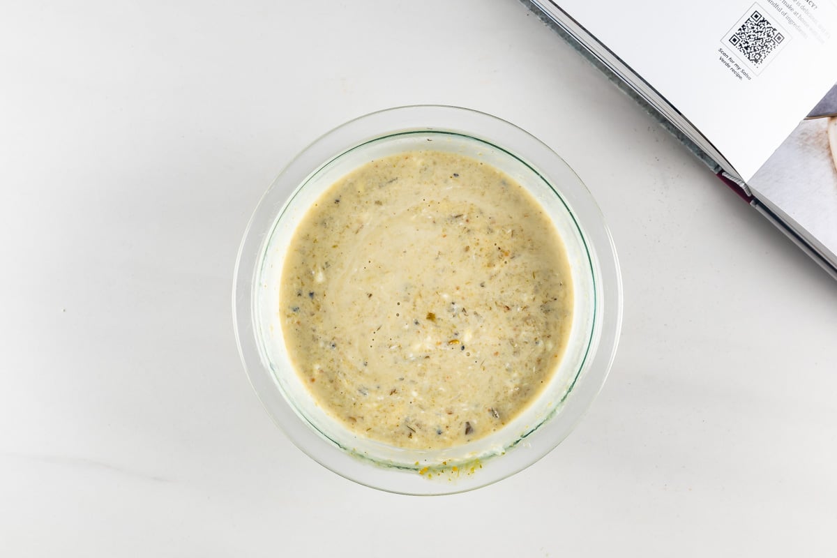 A clear glass bowl filled with a creamy blended soup with green and brown specks. The bowl is placed on a light surface next to an open book. The texture of the soup is smooth, indicating it has been pureed.