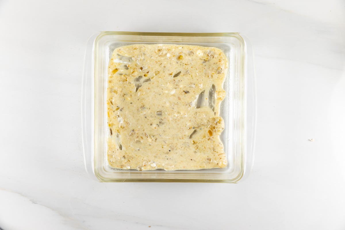 A glass baking dish filled with unbaked yeast batter, featuring visible spices and small pieces of chopped nuts. The dish is placed on a white marble countertop.