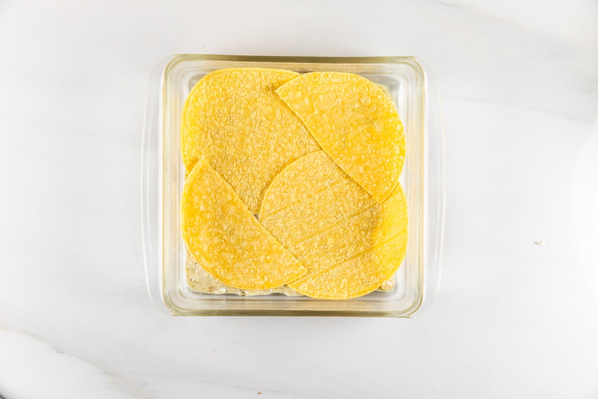 A glass baking dish filled with layered yellow corn tortillas on a white marble surface, ready for assembling a dish.