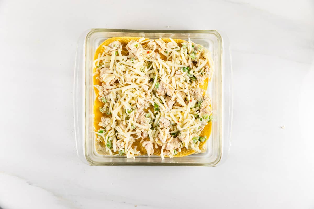 A glass baking dish filled with a mixture of shredded cheese, chopped cooked chicken, and vegetables, placed on a white marble countertop.