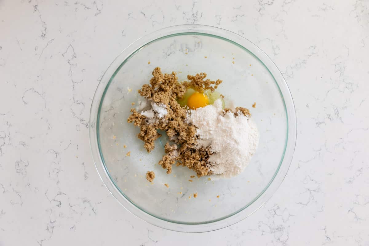 A glass bowl on a marble countertop contains brown sugar, a cracked egg, flour, and green chopped apples. The ingredients are ready to be mixed for baking.