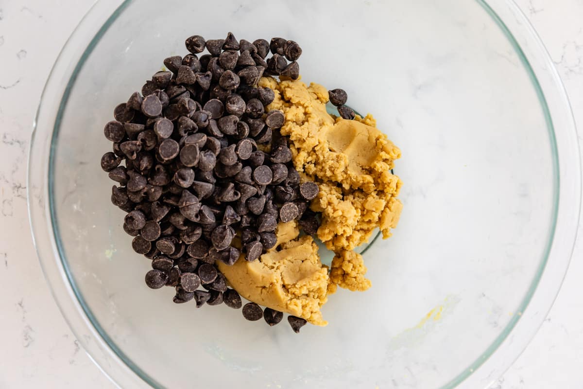 A glass bowl containing chunks of cookie dough and dark chocolate chips on a light countertop.