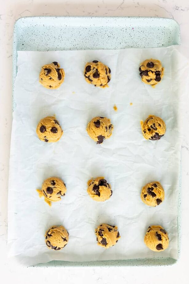 A baking tray with parchment paper holds twelve evenly spaced raw chocolate chip cookie dough balls, ready to be baked. The tray is placed on a white marble countertop.