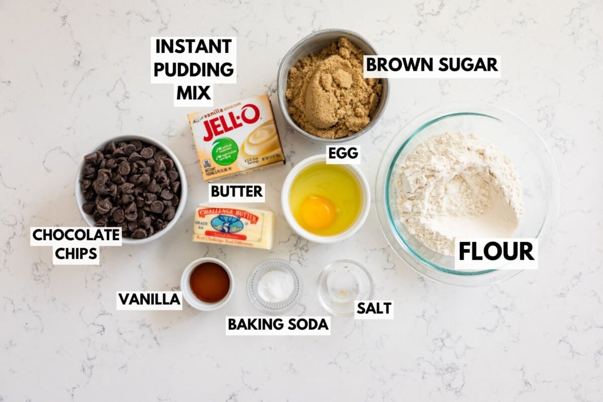 Ingredients for baking: a bowl of chocolate chips, a package of instant pudding mix, brown sugar, a stick of butter, an egg, flour, vanilla, baking soda, and salt, all arranged on a countertop.