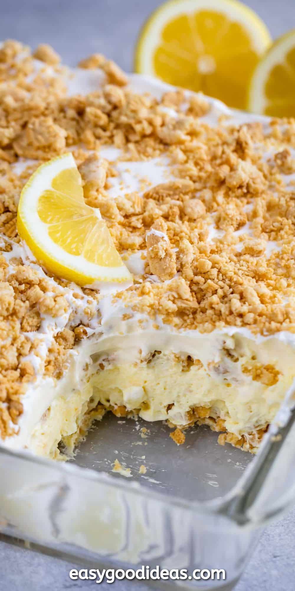 A close-up of a creamy lemon dessert in a glass dish. The dessert is topped with crumbled cookies and a slice of lemon. A portion is missing from the dish, revealing its layers. Two whole lemons are in the background.