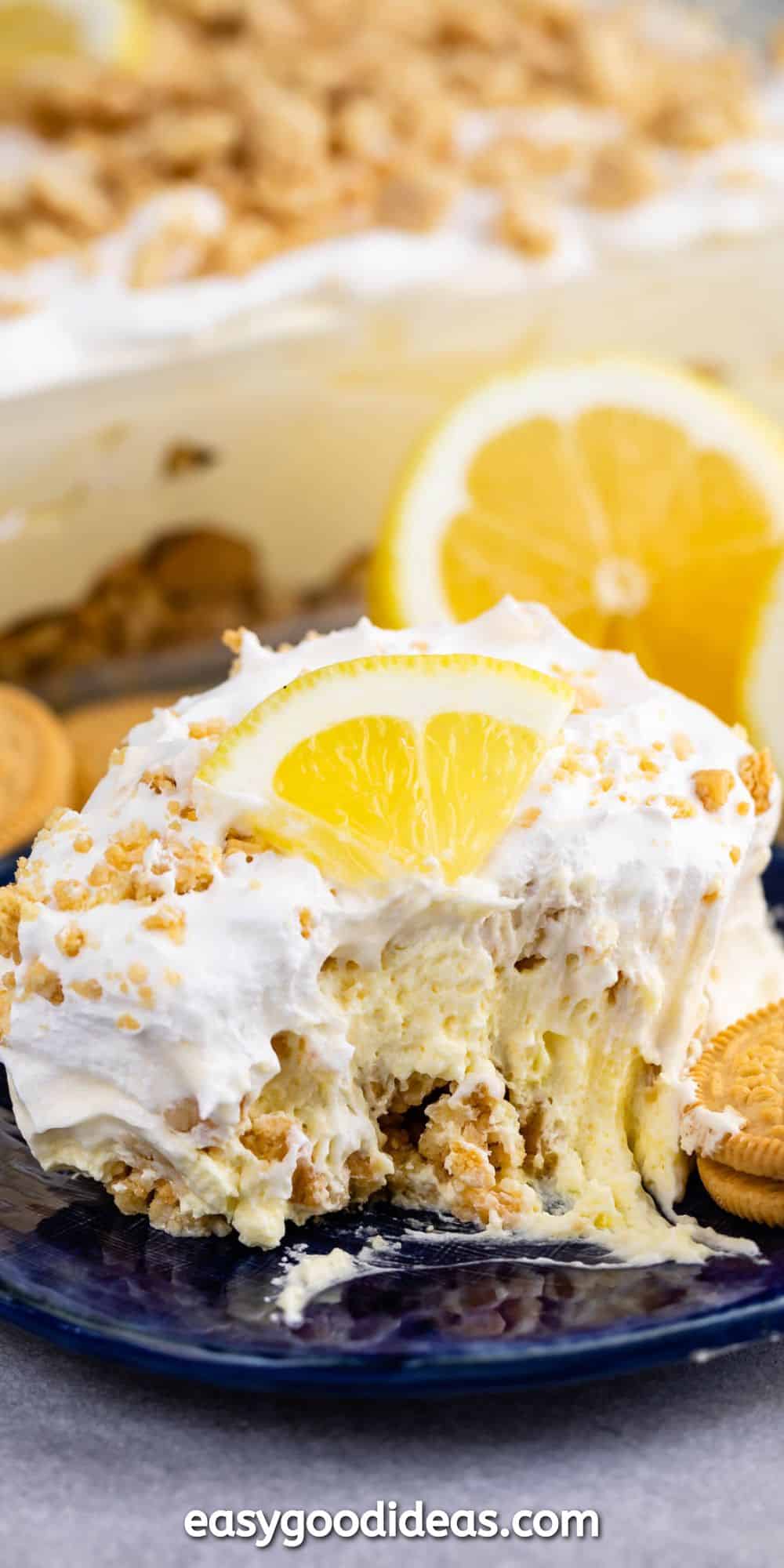 A slice of creamy dessert topped with crumbled cookies and a lemon wedge on a dark plate. In the background, the rest of the dessert sits in a dish, surrounded by whole lemons and cookies.