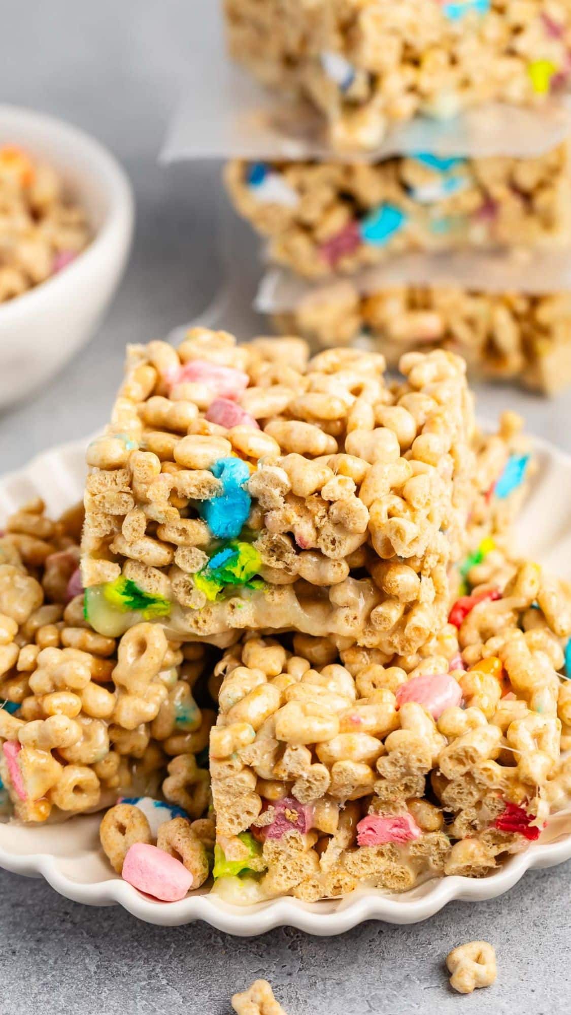 Close-up of Rice Krispie treats mixed with Lucky Charms cereal, stacked on a white plate. The colorful marshmallows and cereal bits are prominent, creating a playful and appetizing appearance.