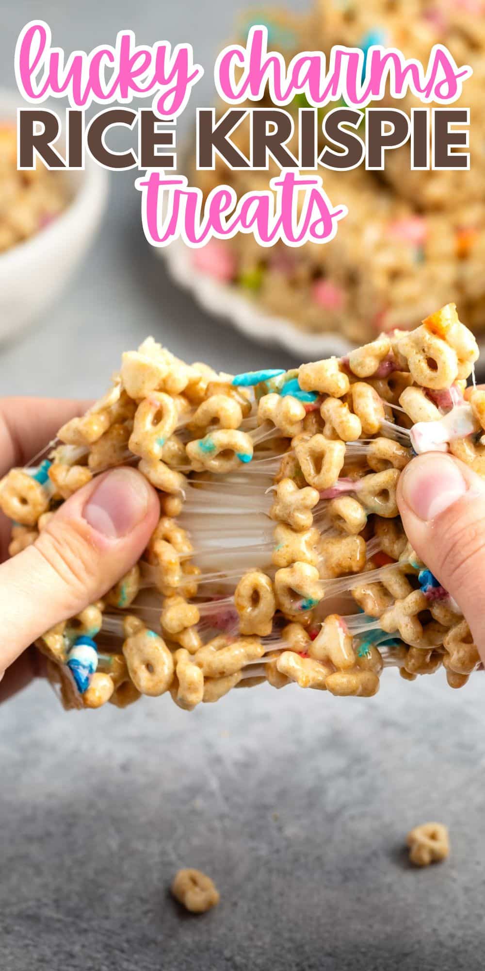 Close-up of hands pulling apart a Lucky Charms Rice Krispie treat, showing its gooey, marshmallowy texture. The treat is packed with colorful cereal pieces. The text above reads, Lucky Charms Rice Krispie Treats.