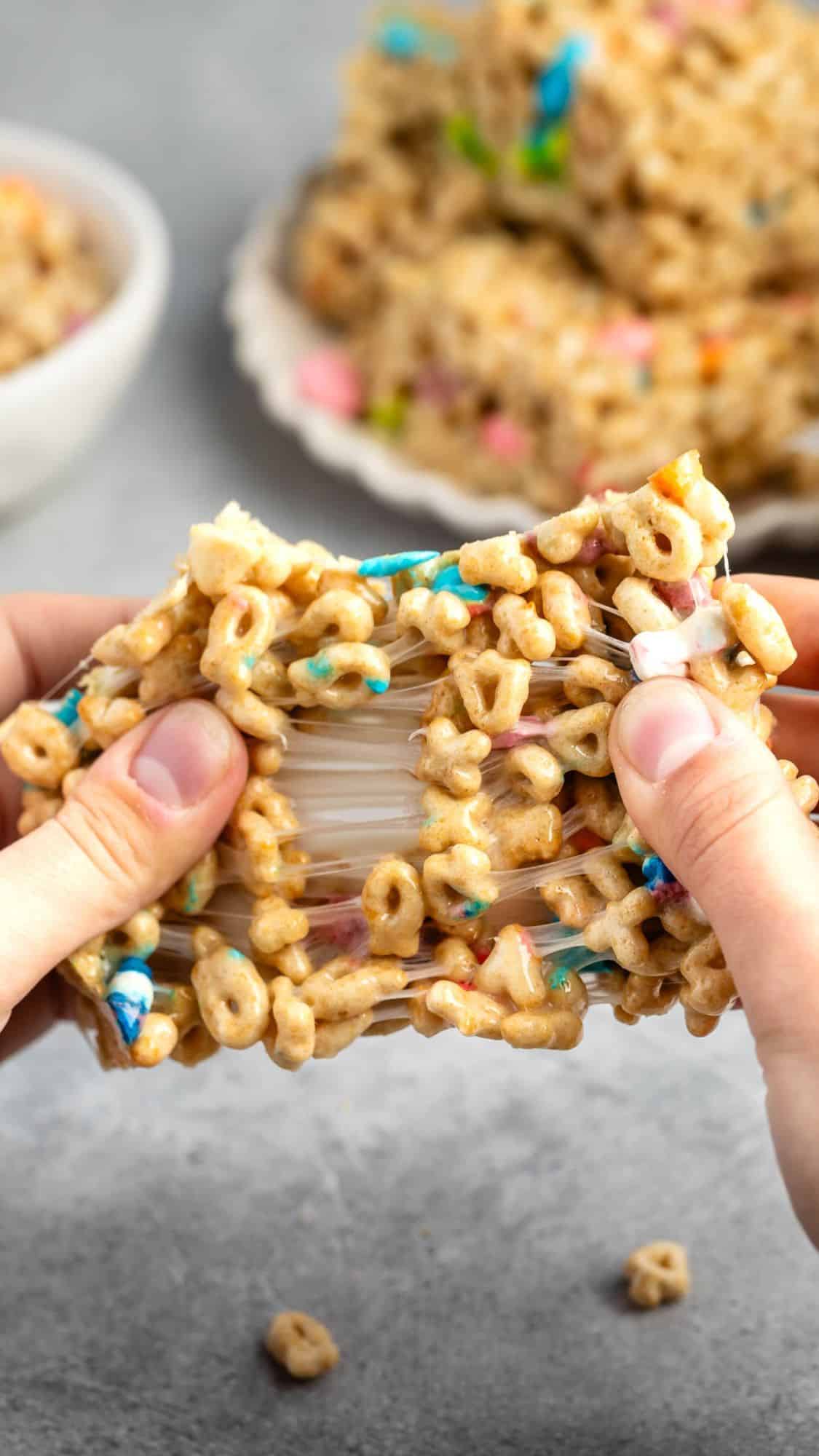 Close-up of hands pulling apart a Lucky Charms Rice Krispie treat, showing its gooey, marshmallowy texture. The treat is packed with colorful cereal pieces.