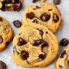 Close-up of freshly baked chocolate chip cookies with golden edges and melty chocolate pieces on parchment paper. Scattered chocolate chips are visible around the cookies.