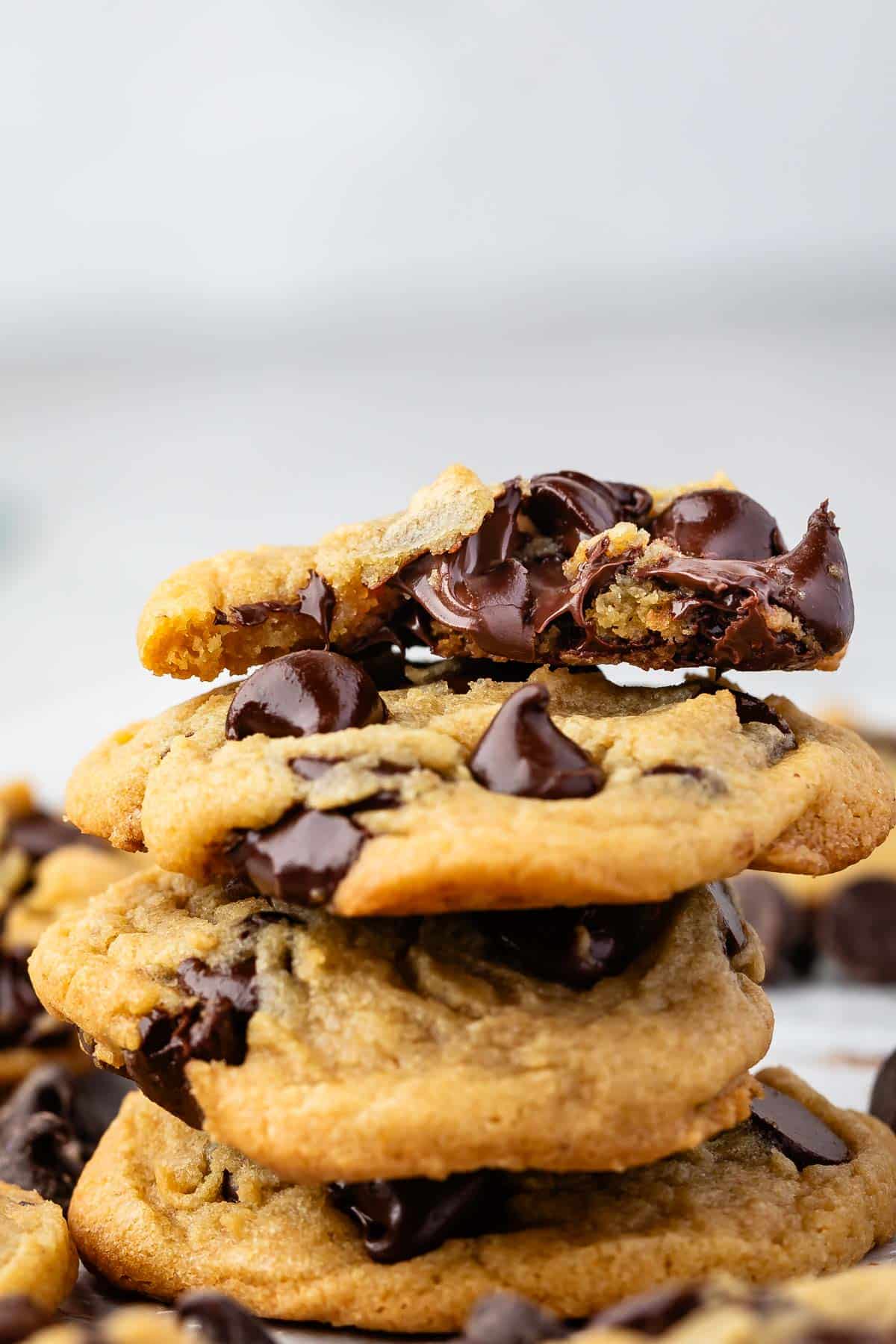 A stack of chocolate chip cookies is shown, with gooey melted chocolate oozing from a broken piece on top. The cookies are golden brown and appear fresh and delicious.