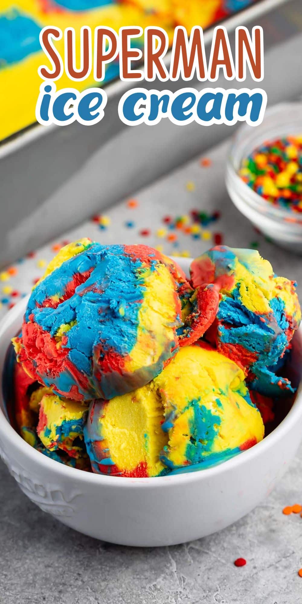 A bowl of vibrant Superman ice cream with swirls of red, blue, and yellow, accompanied by scattered sprinkles on a gray surface.