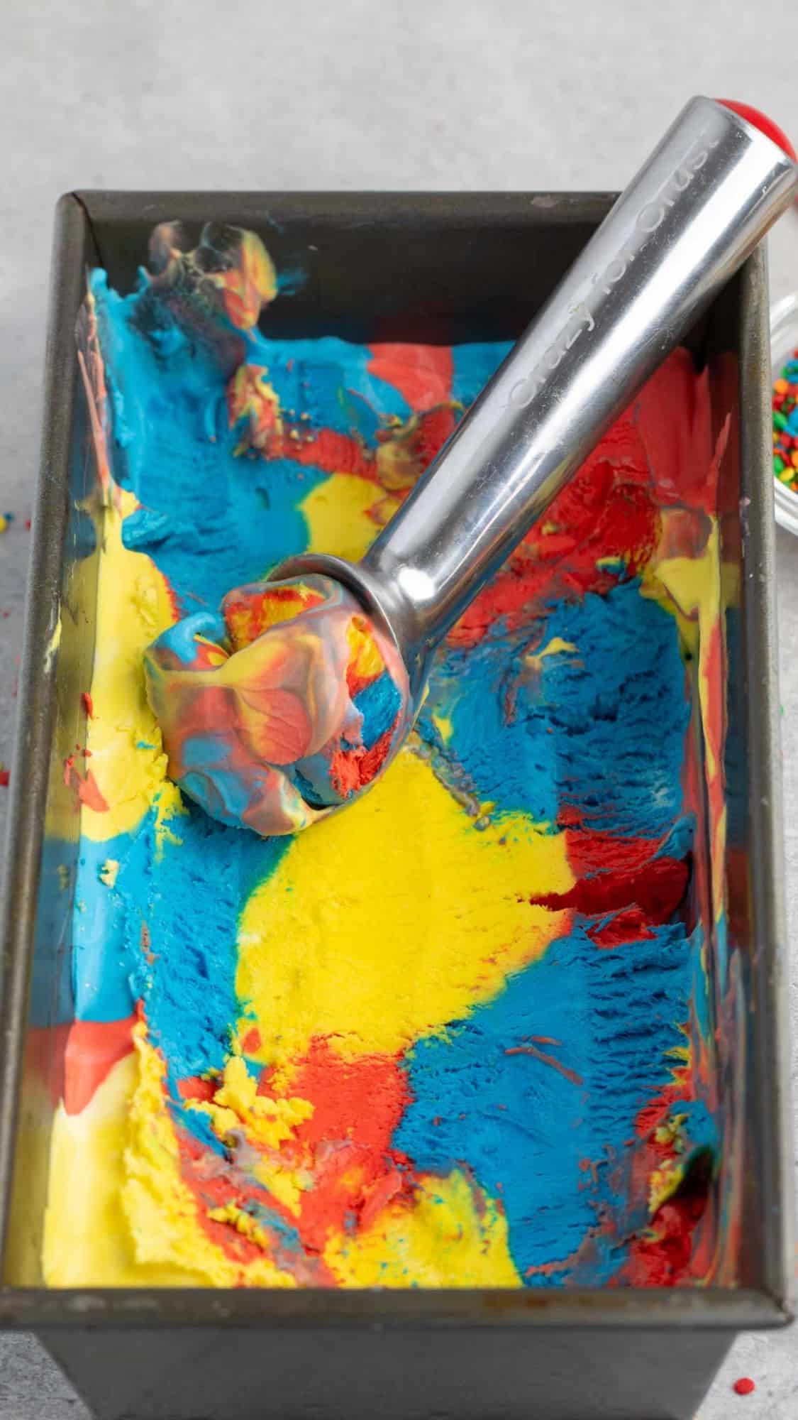 A rectangular metal container holding colorful, swirled ice cream in shades of blue, yellow, and red, with an ice cream scoop resting inside. The background is a light grey surface.