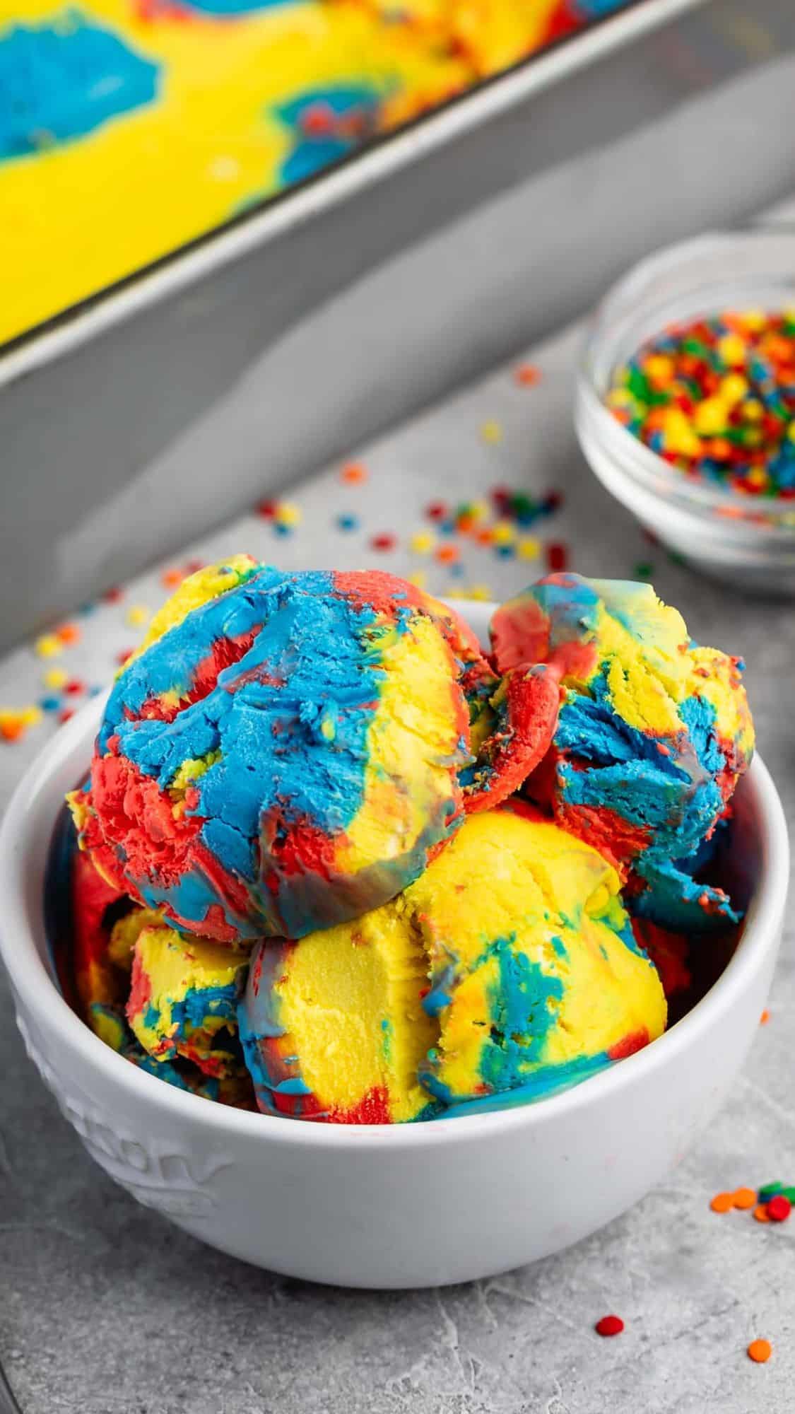 A bowl of colorful ice cream scoops in vibrant shades of blue, red, and yellow. The background includes a container with more ice cream and a small bowl of multicolored sprinkles, all on a gray surface.