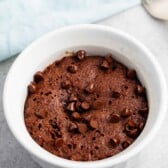 A chocolate mug cake topped with chocolate chips in a white ramekin, placed on a gray surface. A silver fork is partially visible in the background, and a light blue cloth is nearby.